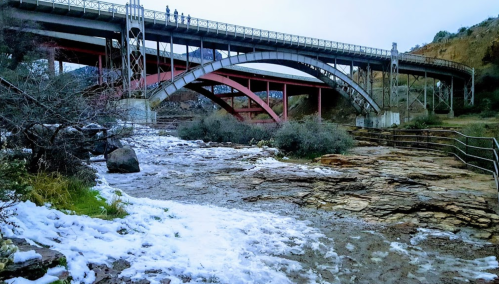 A bridge arches over a rocky riverbank with foam on the water, surrounded by greenery and a cloudy sky.